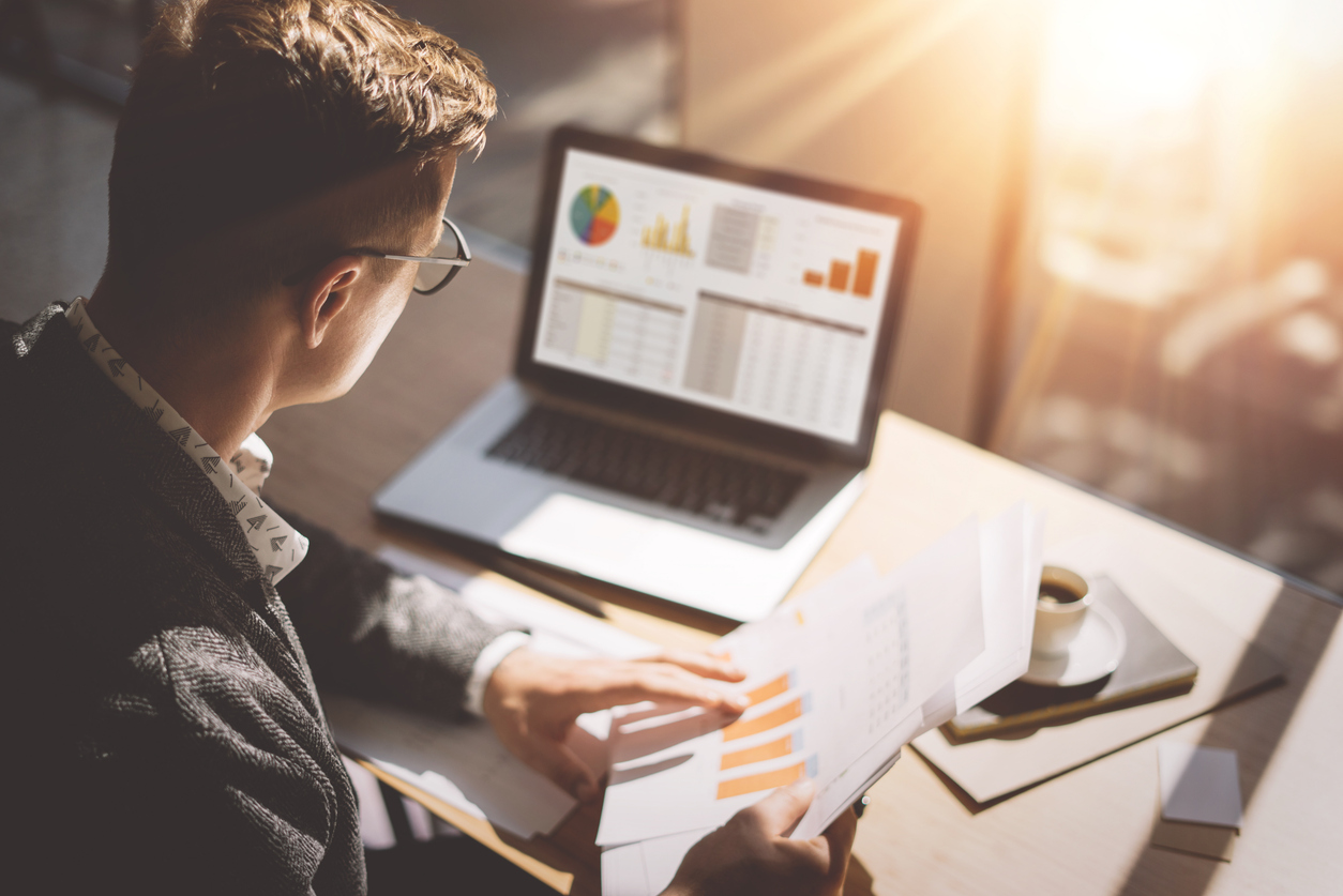 young finance market analyst in eyeglasses working at sunny office on laptop while sitting at wooden table.businessman analyze document in his hands.graphs and diagramm on notebook screen.blurred.